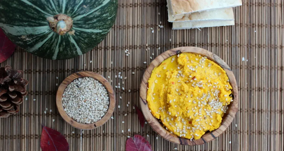 Ein leuchtend gelber Hummus, bestreut mit Sesam, wartet neben einer Schale voller Sesam und einem Kürbis. Perfekt für ein kleines Hummus-Baby-Festmahl!