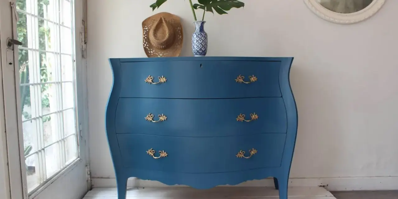 Cómoda antigua con **muebles pintados en azul** y tiradores dorados, decorada con un sombrero y un jarrón con hojas.