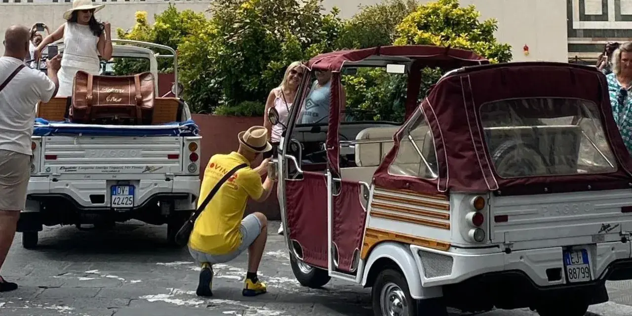 Turisti scattano foto a veicoli atipici, un Piaggio Ape e un tuk-tuk, in una strada soleggiata.