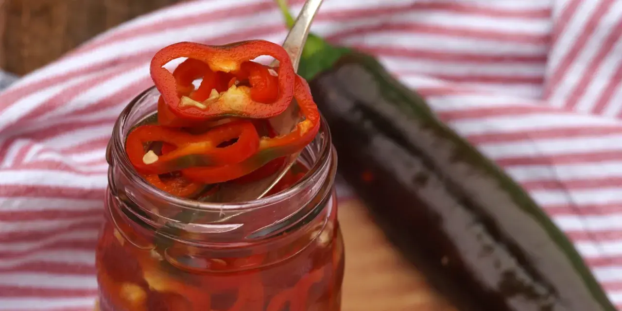 En burk med picklad chili, med en sked som lyfter upp en skiva. En mörk chili ligger bredvid.