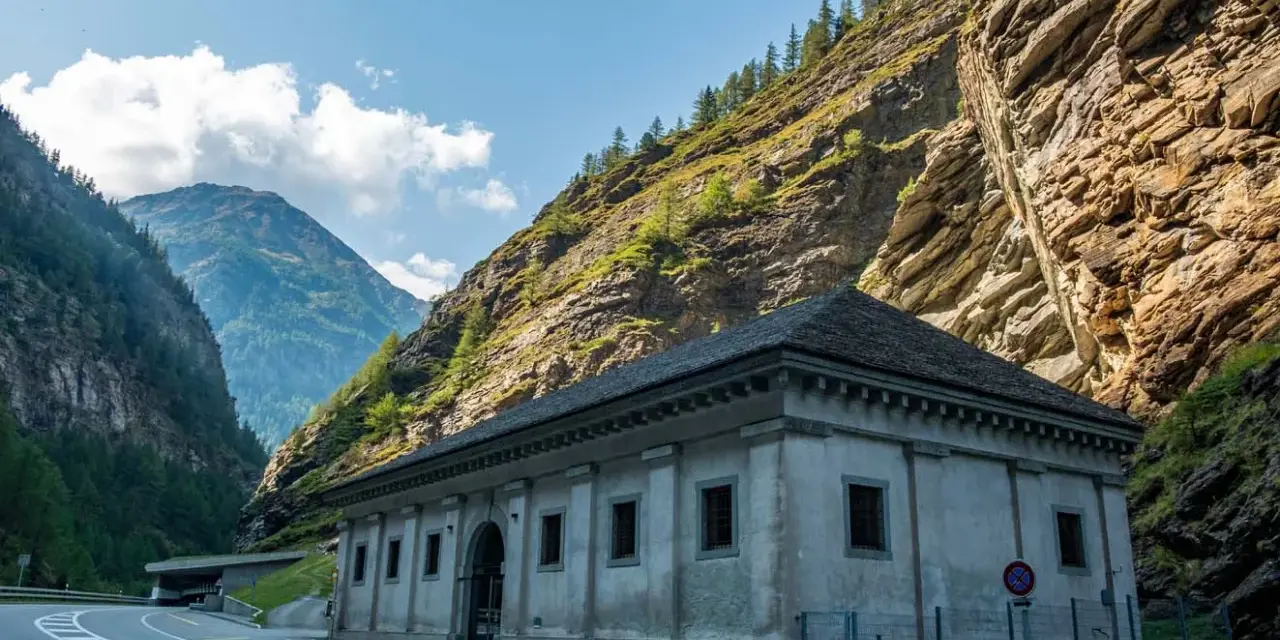 Edificio in pietra ai piedi di una montagna rocciosa, con strada e tunnel. Un passo alpino che sembra non finire mai, un vero sempione passo.