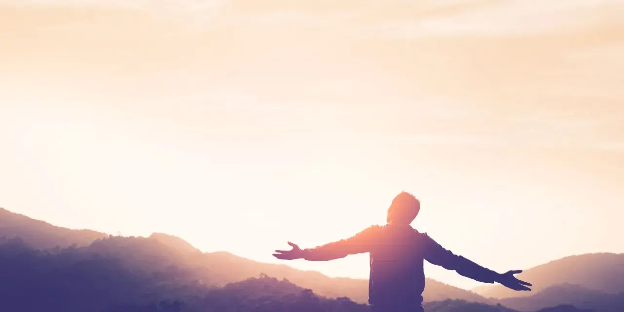 Silhouette einer Person mit ausgebreiteten Armen vor einer Bergkulisse bei Sonnenuntergang. Ein Symbol für Kraft und Hoffnung nach einer Krankheit.