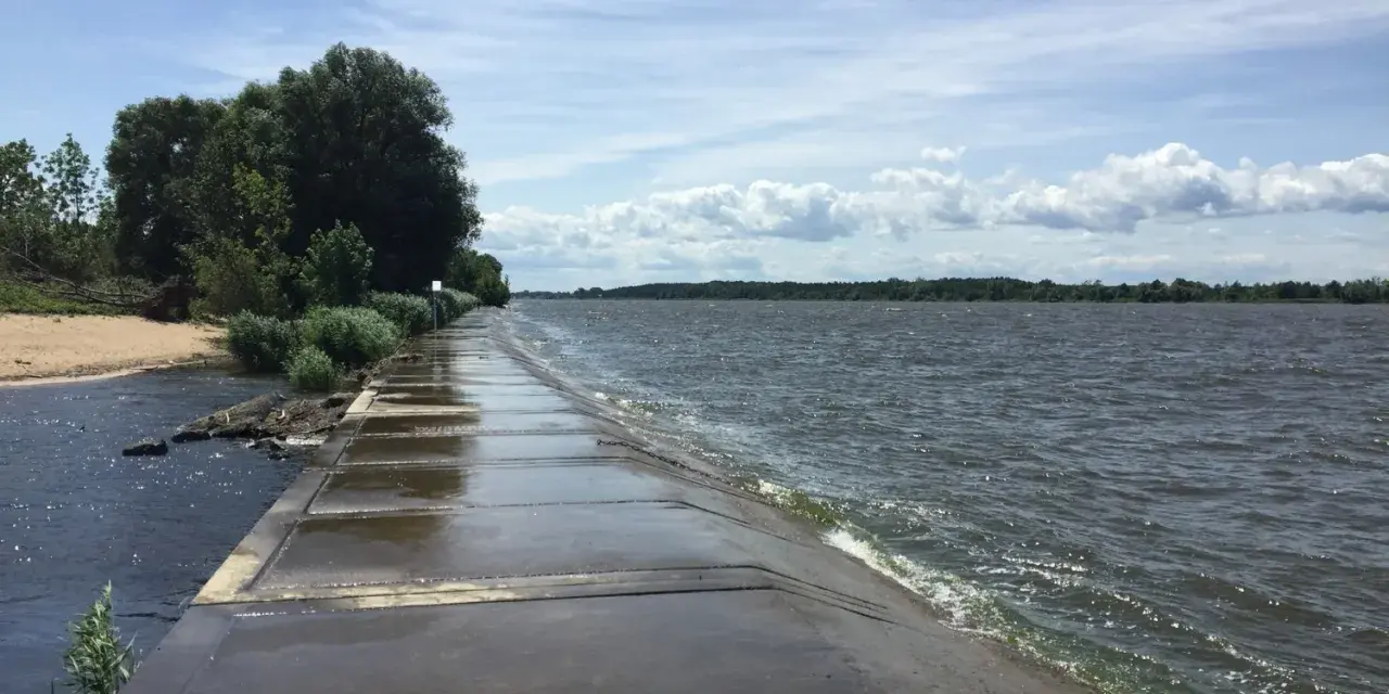 Betonowy falochron w miejscu ujścia Wisły. Woda faluje, a po lewej stronie widać piaszczystą plażę i zieleń.