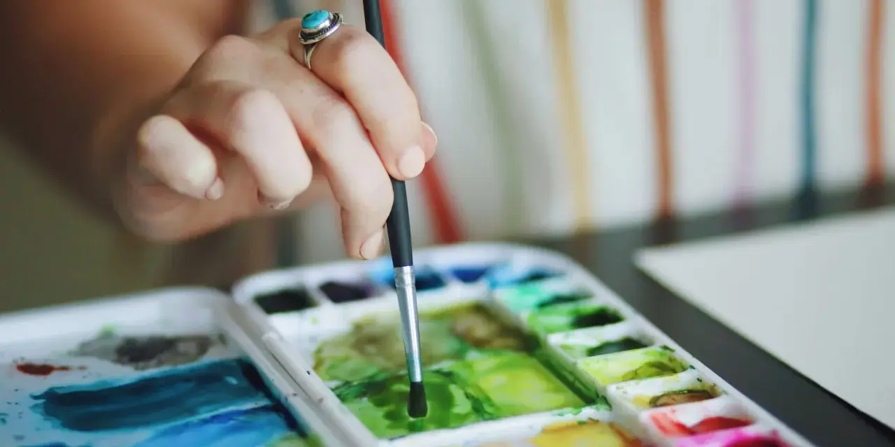 Main d'une artiste, bague turquoise, tenant un pinceau au-dessus d'une palette de couleurs vives de **de l'aquarelle**.