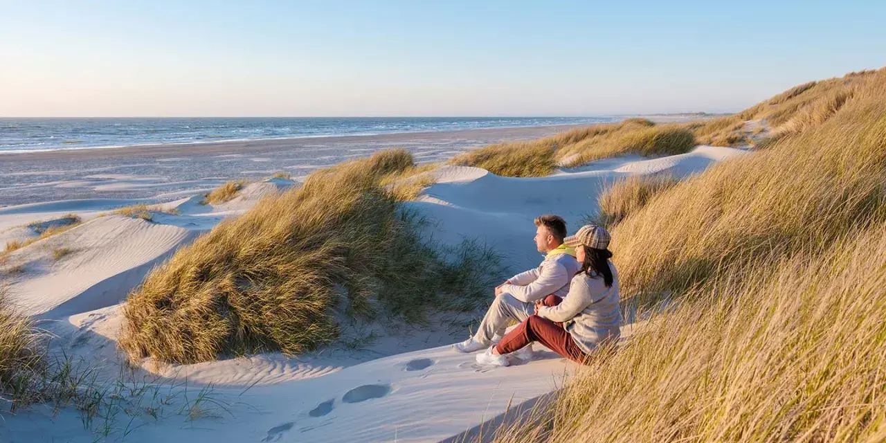 Ett par sitter på en sanddyn och njuter av utsikten över havet. Vackra sanddyner med gräs sträcker sig längs kusten, en idyllisk plats i en nationalpark i Danmark.