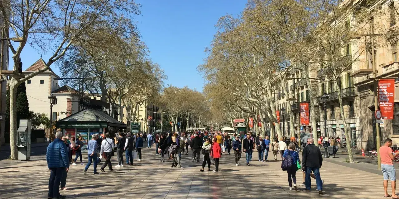 Tłum ludzi spaceruje po słonecznej La Rambla w Barcelonie, otoczonej drzewami i zabytkowymi budynkami.