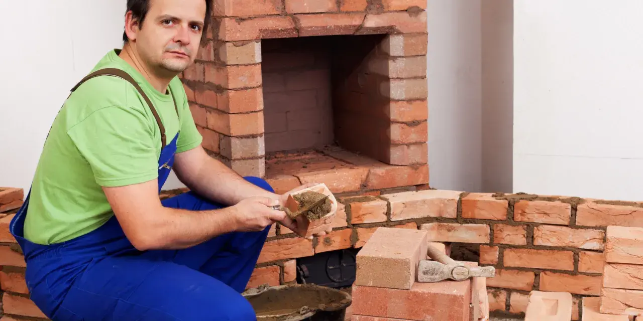 Murarz układa cegły szamotowe, budując kominek. W wiadrze zaprawa, obok stosy cegieł.