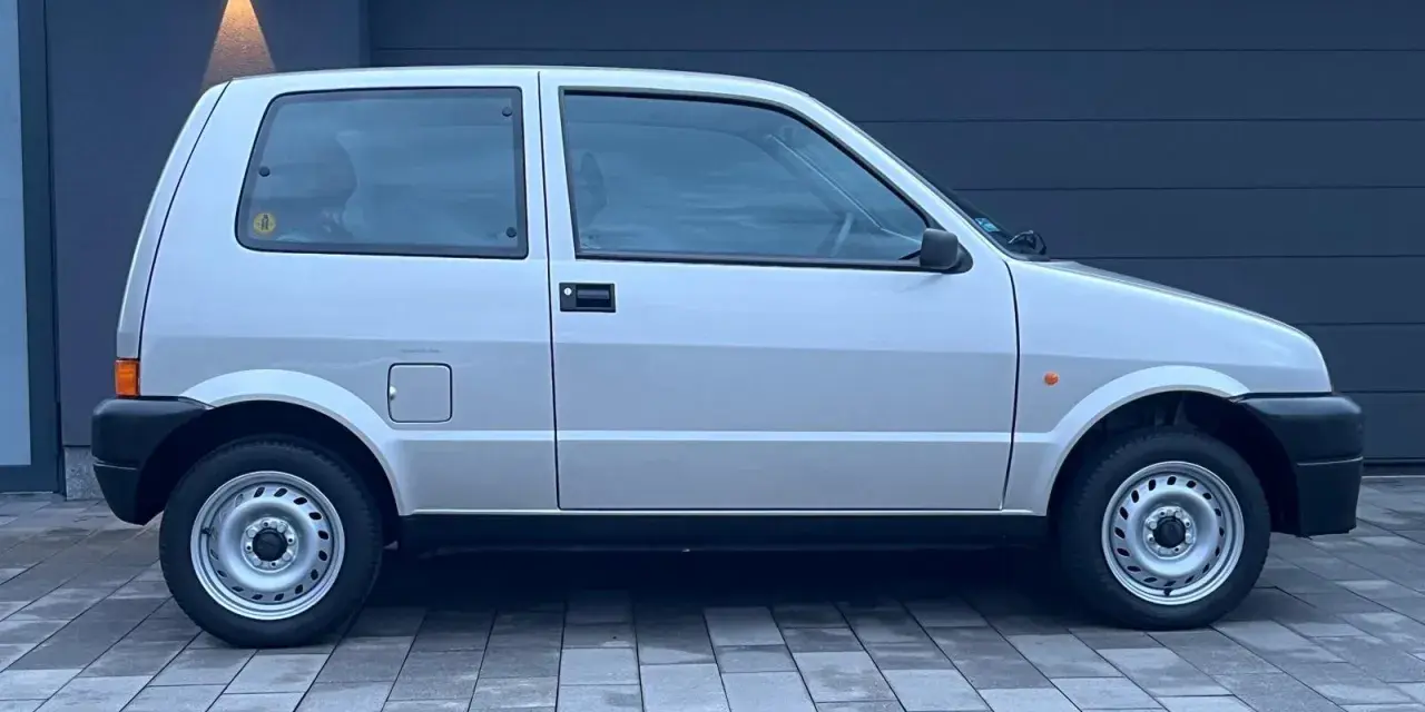 Srebrny Fiat Cinquecento na tle ciemnego garażu, z charakterystycznymi felgami i czarnymi zderzakami.