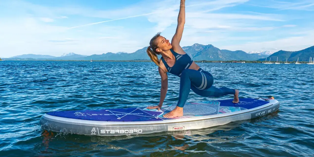 Frau praktiziert Krieger II auf einem SUP-Board auf ruhigem Wasser, Berge im Hintergrund.