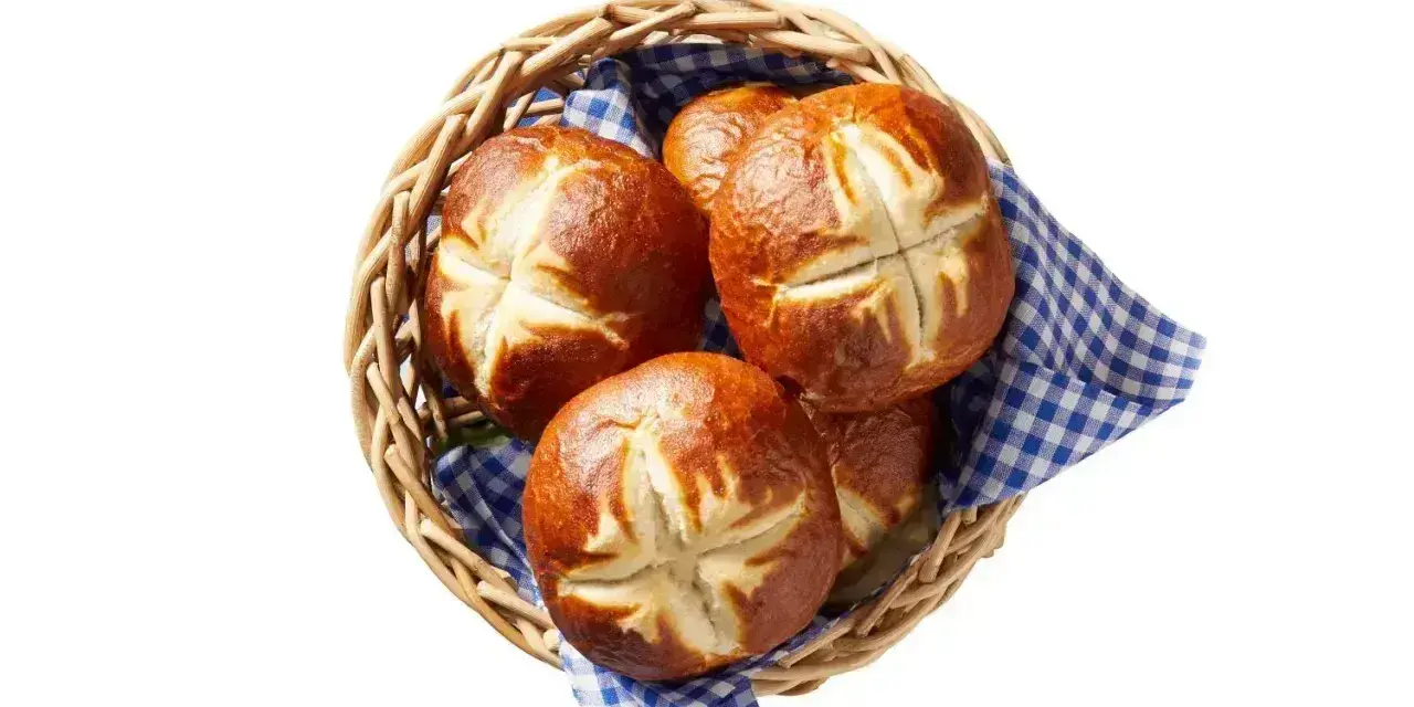 Koszyk z kilkoma bułkami bawarskimi, ułożonymi na niebiesko-białej kratkowanej serwetce.