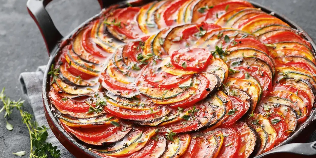 Ein wunderschönes Ratatouille-Rezept, das in einer roten Pfanne mit geschichteten Tomaten-, Zucchini- und Auberginenscheiben und frischen Kräutern präsentiert wird.