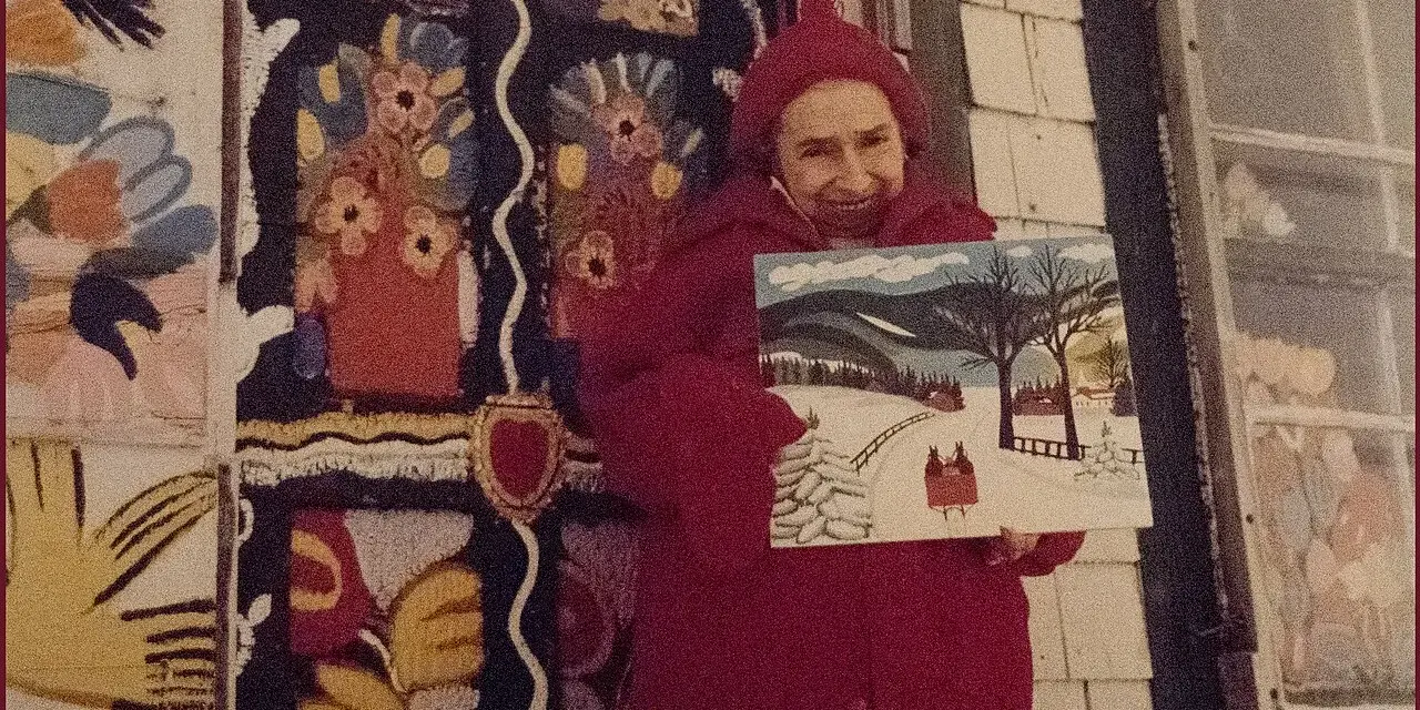 Maud Lewis, klädd i rött, håller upp en av sina charmiga vintermålningar framför sitt färgstarka hem.