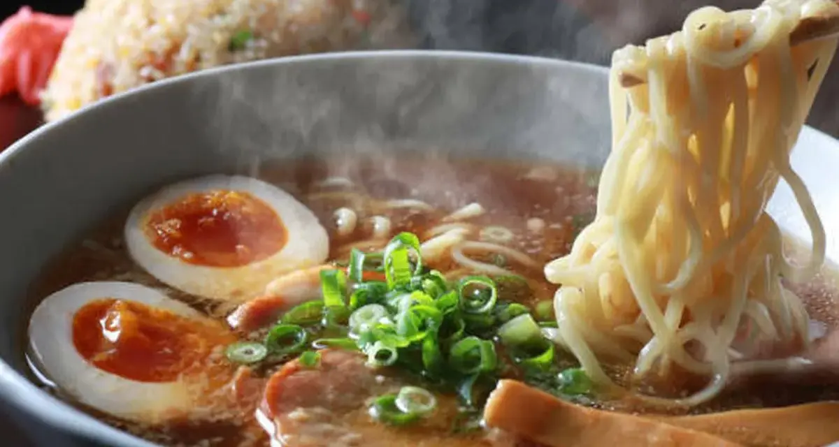 Un humeante tazón de ramen con huevo cocido, cebolleta y cerdo. Los palillos levantan fideos, listos para disfrutar de esta deliciosa receta ramen.