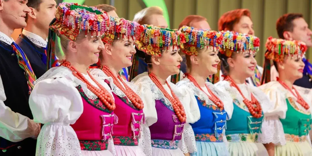 Szła dzieweczka w tradycyjnym stroju, z barwnym wiankiem na głowie, obok innych w ludowych strojach.