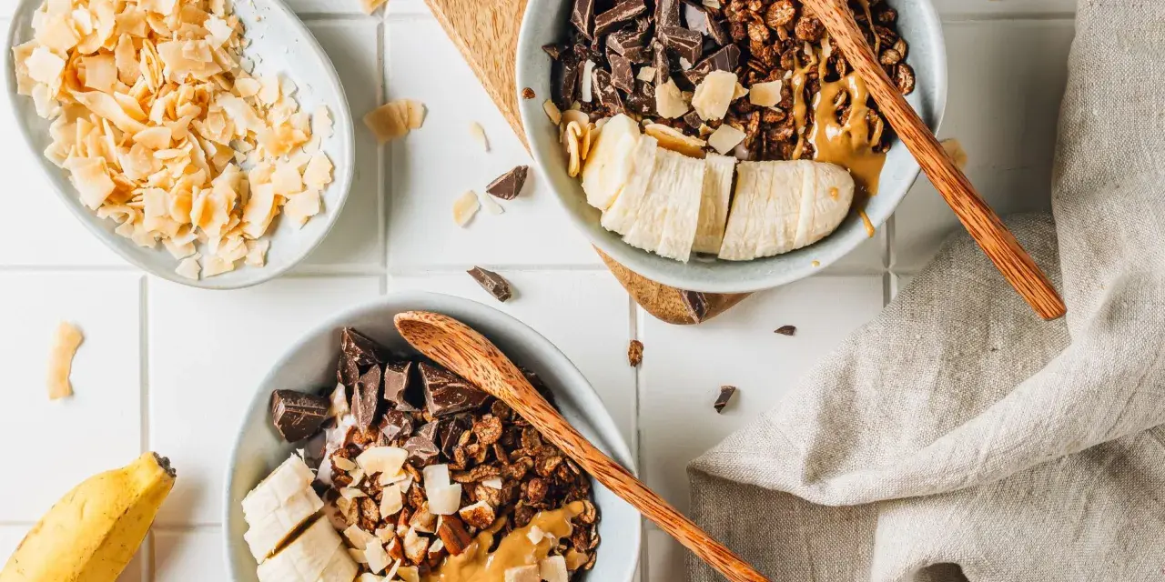 Due ciotole con granola fatta in casa, banana a fette, cioccolato fondente e burro d'arachidi. Cocco in scaglie a lato.