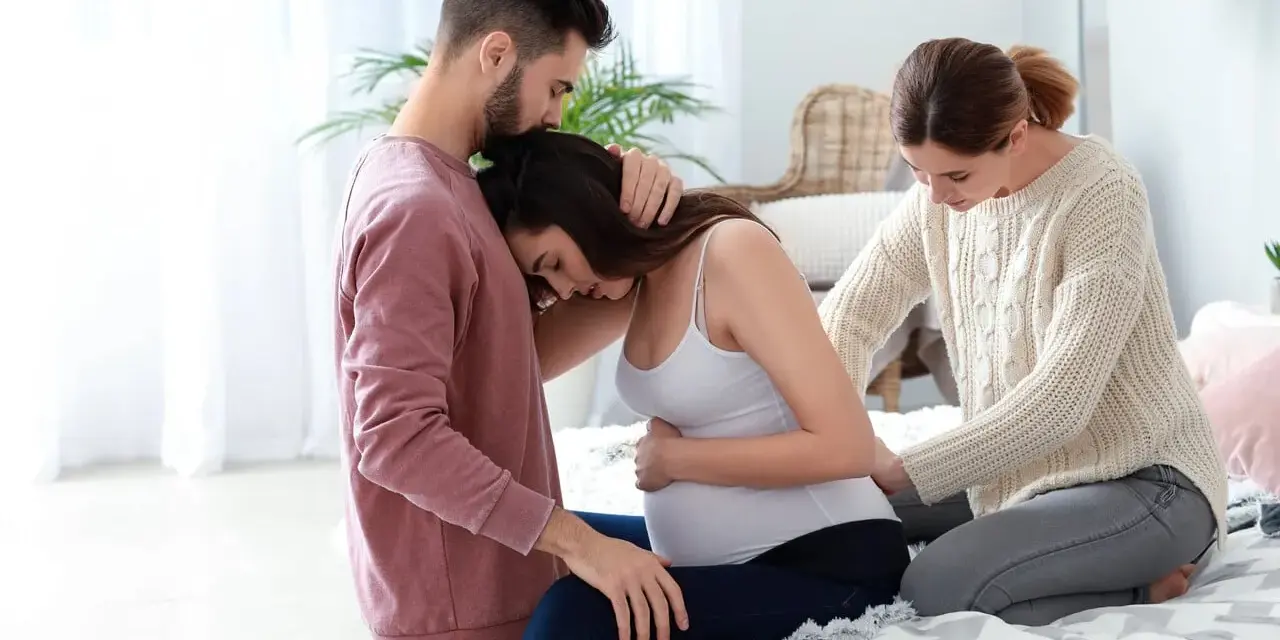 Mężczyzna wspiera ciężarną kobietę, a doula delikatnie masuje jej plecy.