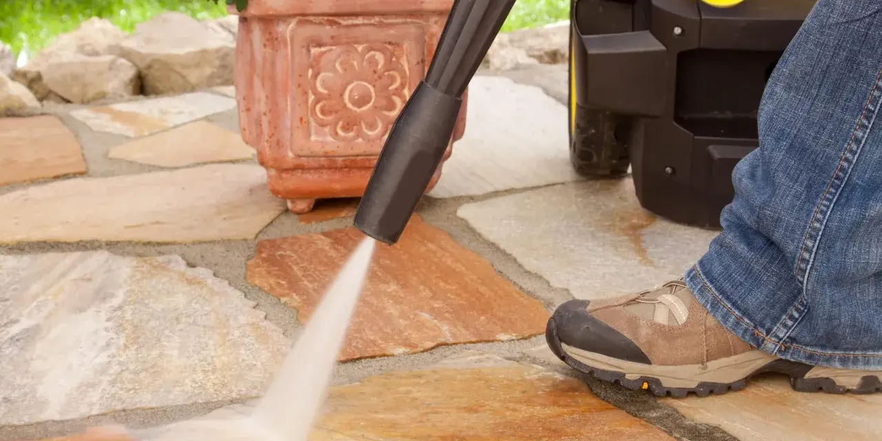 Pulire pavimento esterno con idropulitrice. Un getto d'acqua potente rimuove lo sporco dalle pietre del patio, mentre una persona in jeans e scarponi osserva.
