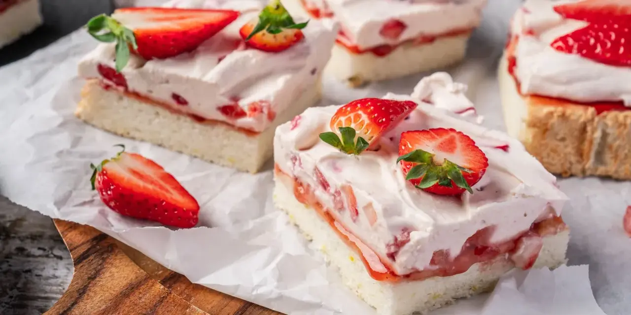 Saftige Stücke eines Erdbeerkuchen Blechs mit frischen Erdbeeren und Sahne, auf Backpapier angerichtet.