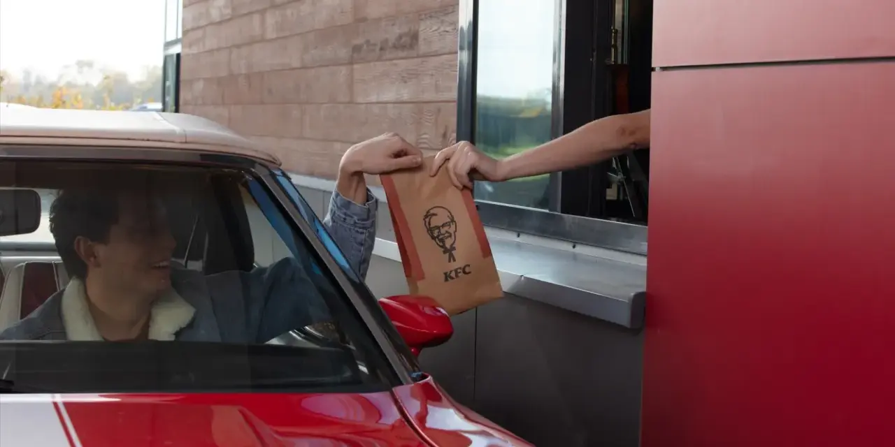 Mężczyzna w czerwonym aucie odbiera z okienka KFC torbę z jedzeniem. Wygląda na zadowolonego, że otrzymał swój mega pocket kcal.