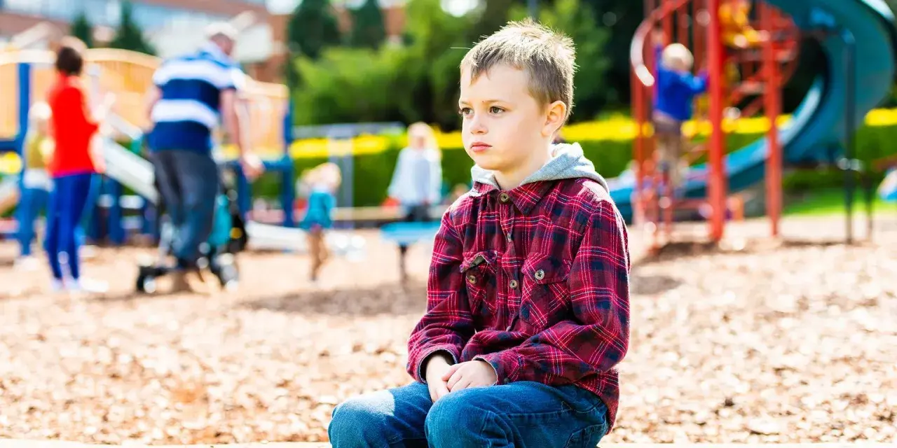 Chłopiec z zespołem Aspergera siedzi samotnie na placu zabaw, podczas gdy inne dzieci się bawią.