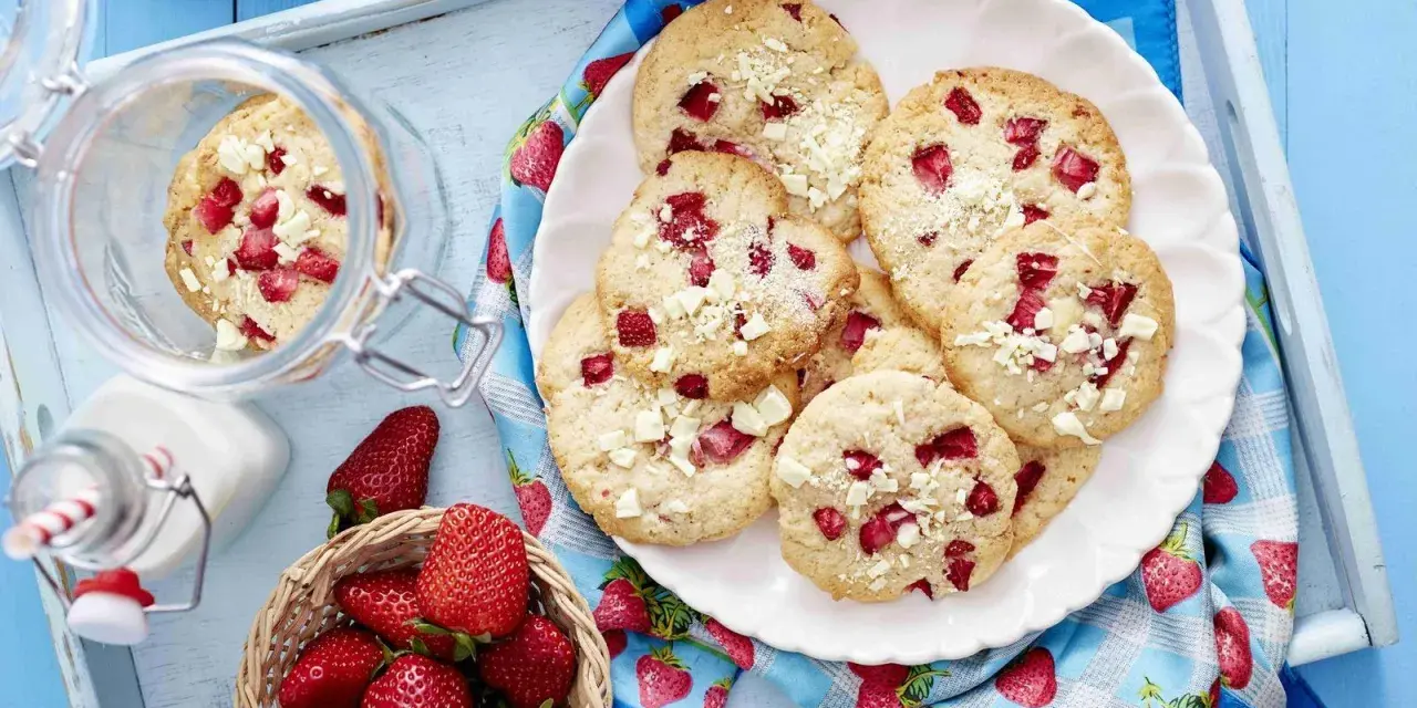 Saftige Erdbeer-Cookies mit weißen Schokostückchen auf einem Teller, daneben frische Erdbeeren und ein Glas Milch.