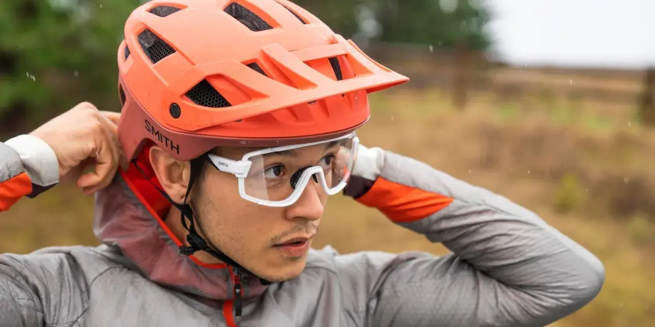 Uomo con casco da bici arancione e occhiali bianchi, mentre regola il casco. Utile per capire come misurare la testa per il casco.