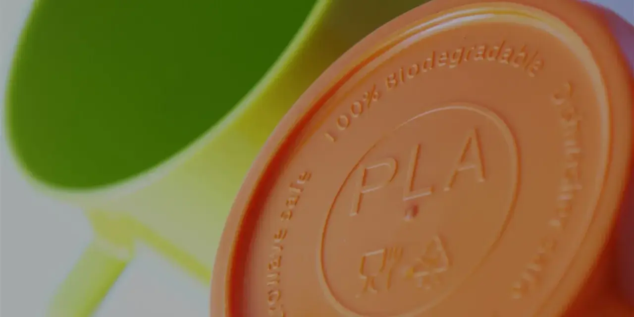 Pomarańczowa pokrywka z napisem PLA, 100% biodegradowalna, obok zielonego kubka.