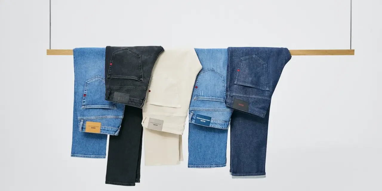 Cinco pares de pantalones vaqueros de distintos tonos cuelgan de una barra de madera. El **denim material** se presenta en azul claro, negro, beige, azul medio y azul oscuro.