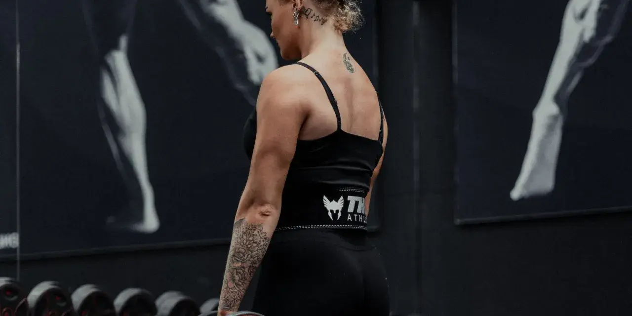 Kvinna med tatueringar utför shrugs med hantlar i ett gym.