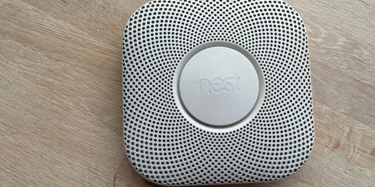 En vit Google Nest Protect-rökdetektor med ett runt "nest"-märke i mitten, placerad på ett trägolv.
