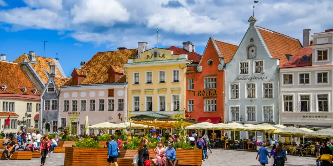 Kolorowe kamienice na placu w Tallinie, stare miasto tętni życiem. Ludzie spacerują, siedzą w kawiarniach pod żółtymi parasolami.