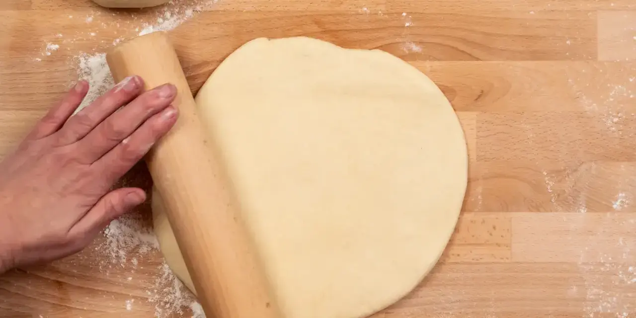 Hand rollt Teig auf Holzbrett aus. Vielleicht mit Dinkelmehl? Was ist Dinkelmehl?