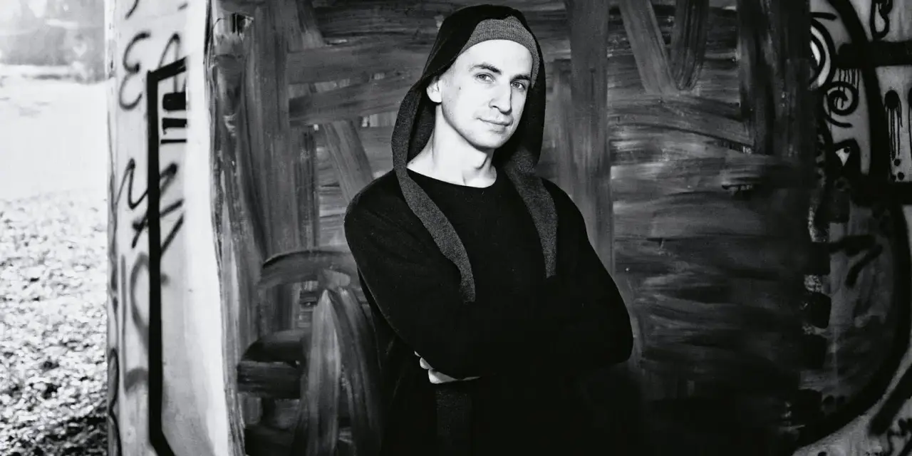 Marek Pędziwiatr w czarnym kapturze, z rękami skrzyżowanymi na piersi, stoi na tle graffiti.