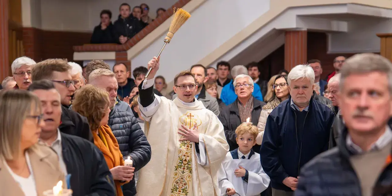 Kapłan błogosławi wiernych podczas liturgii Wielkiej Soboty, unosząc kropidło. W tłumie widać ludzi z zapalonymi świecami.