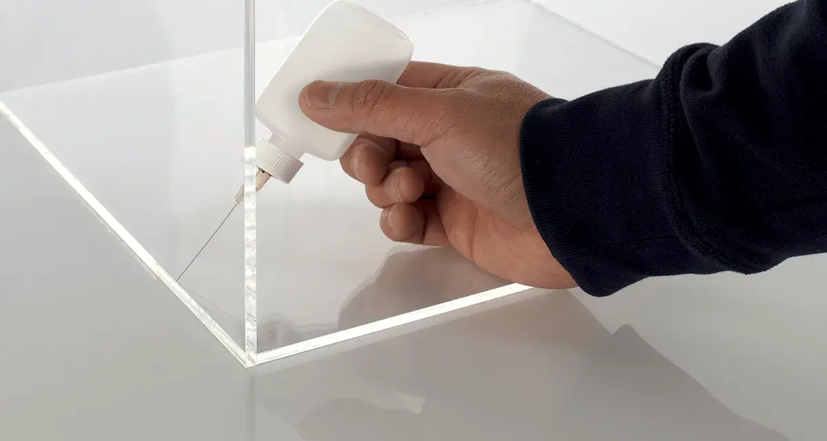 A hand applies adhesive to the corner of a clear plastic box, demonstrating precise gluing of plastic for a clean finish.