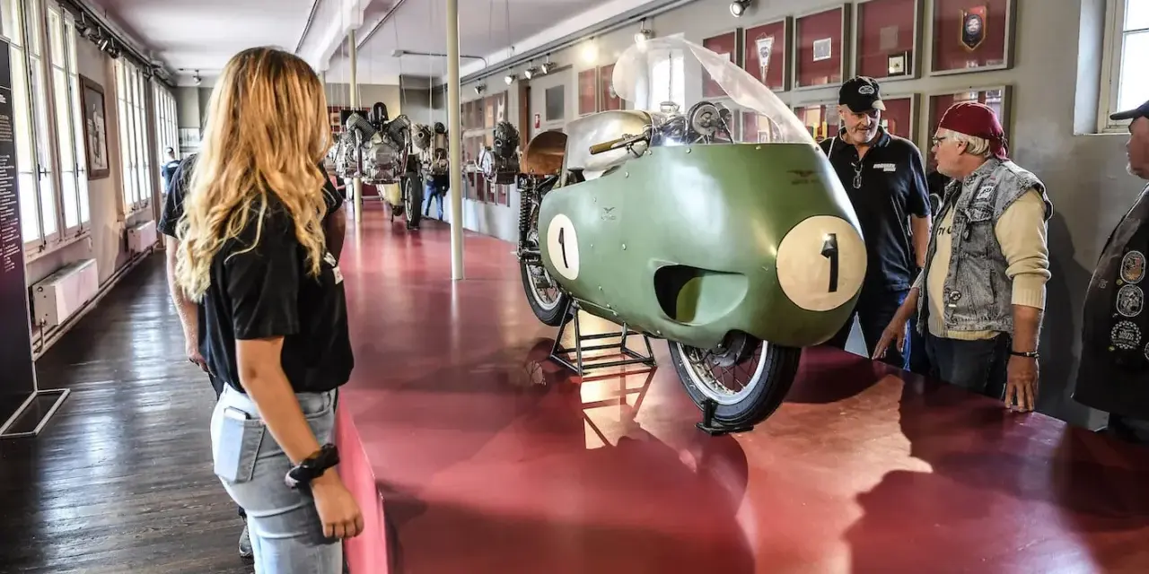 Un raduno moto Guzzi in un museo. Una moto da corsa verde con il numero 1 in primo piano, ammirata da visitatori.