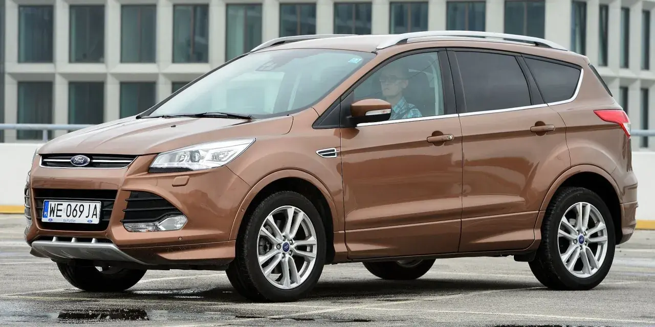 Brązowy Ford Kuga 2019 z kierowcą w okularach, zaparkowany na mokrym parkingu przed nowoczesnym budynkiem.