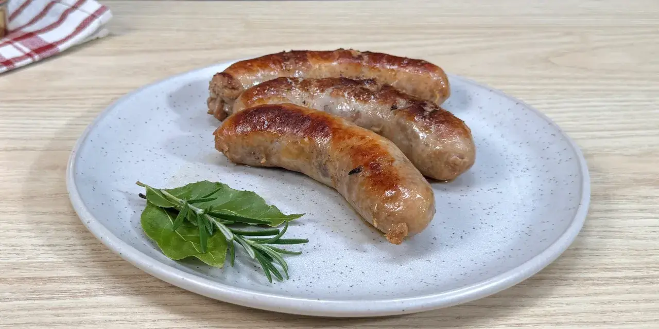 Tre salsicce dorate e succulente, appena cotte in padella, riposano su un piatto con rametti di rosmarino e alloro.
