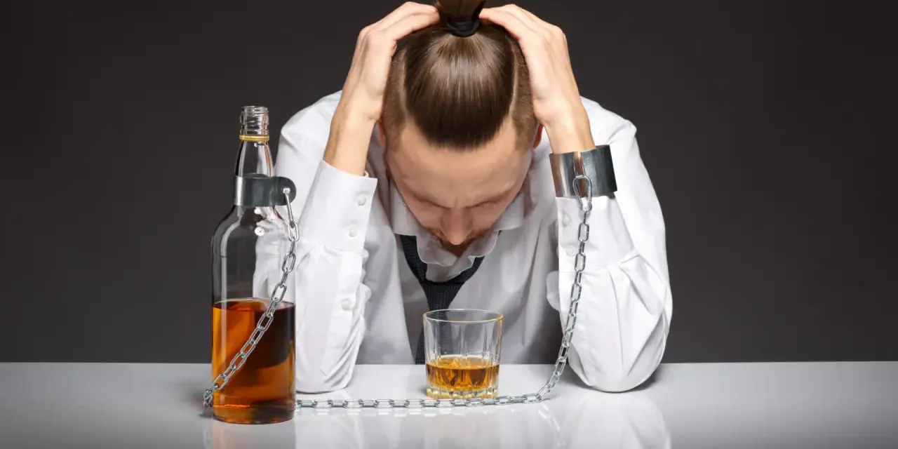 Mężczyzna w kajdanach, przykuty łańcuchem do butelki i szklanki z alkoholem. To symbol zniewolenia przez grupę alkoholową.