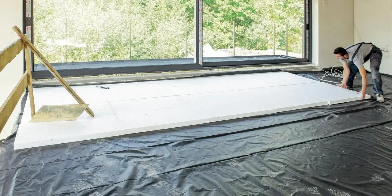 Pracownik układa płyty styropianowe do ocieplenie stropu betonowego. Duże okno wychodzi na zieleń.