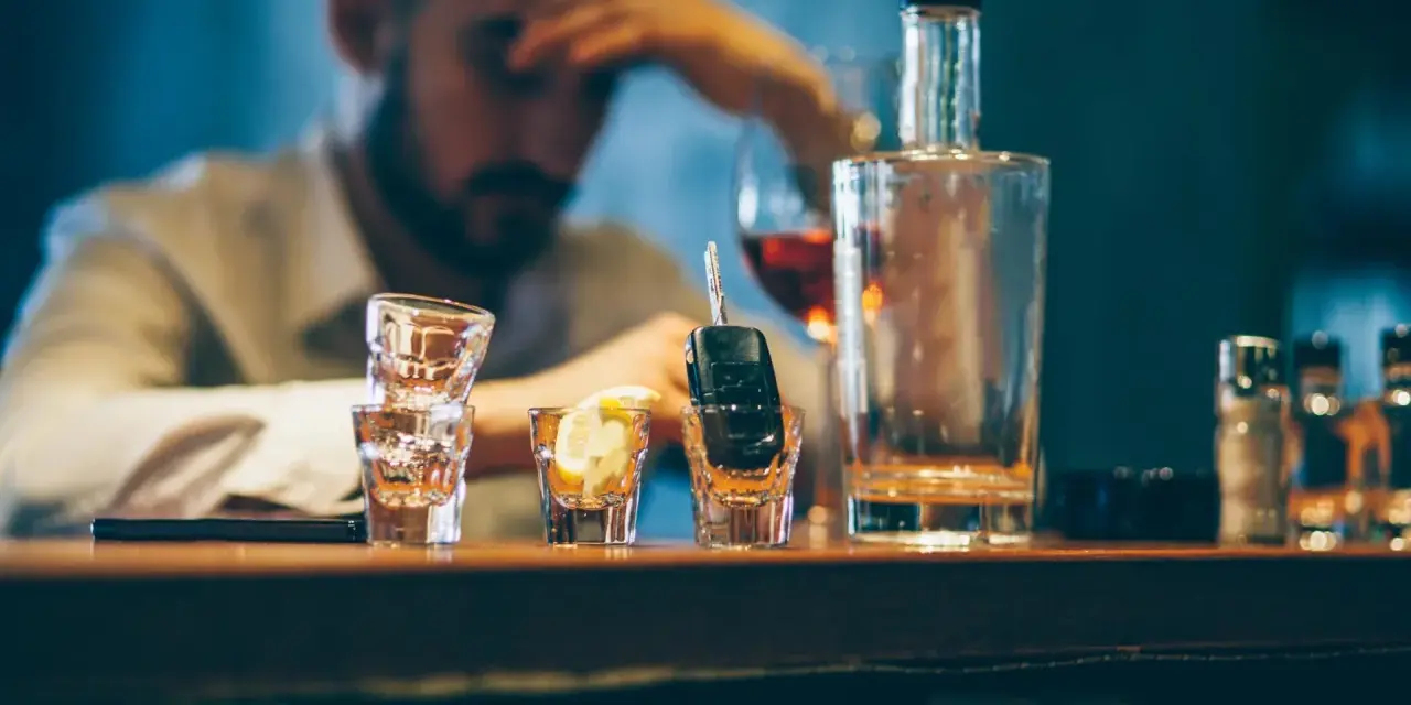 Ein Mann mit Kopfschmerzen sitzt am Tresen, neben Autoschlüsseln in einem Schnapsglas. Alkohol am Steuer unter 21 ist eine Gefahr.