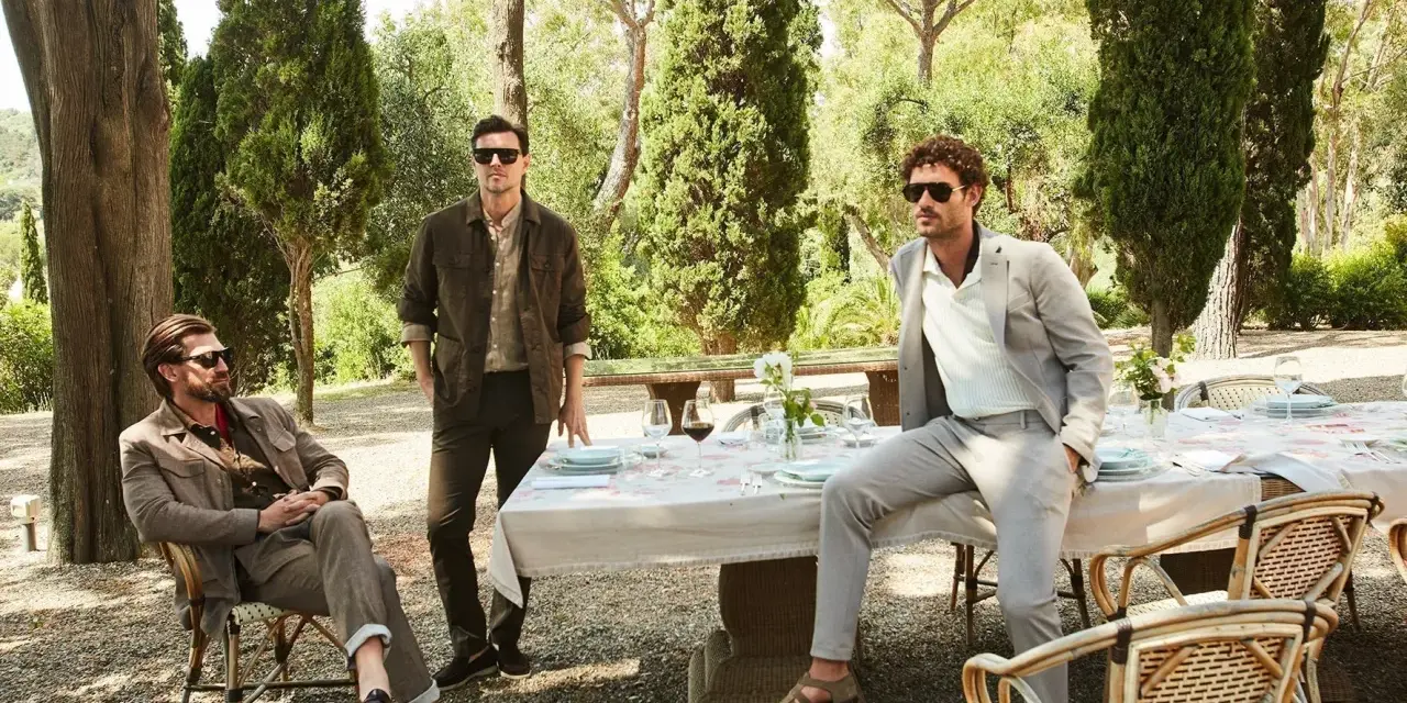 Uomini eleganti in un giardino, con un uomo seduto che indossa un completo marrone e un altro in piedi con un completo grigio. Il pantalone, in entrambi i casi, definisce lo stile.