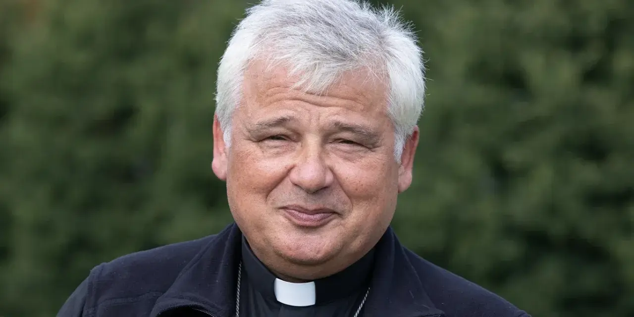 Uśmiechnięty jałmużnik papieski w sutannie i czarnym polarze, na tle zieleni.
