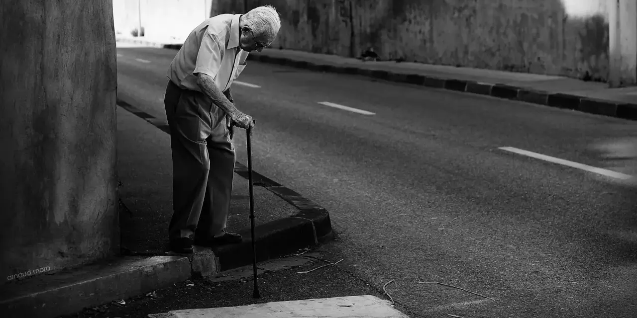 Une photo dans la rue : un vieil homme se penche sur sa canne, le regard perdu, au bord d'une route déserte.