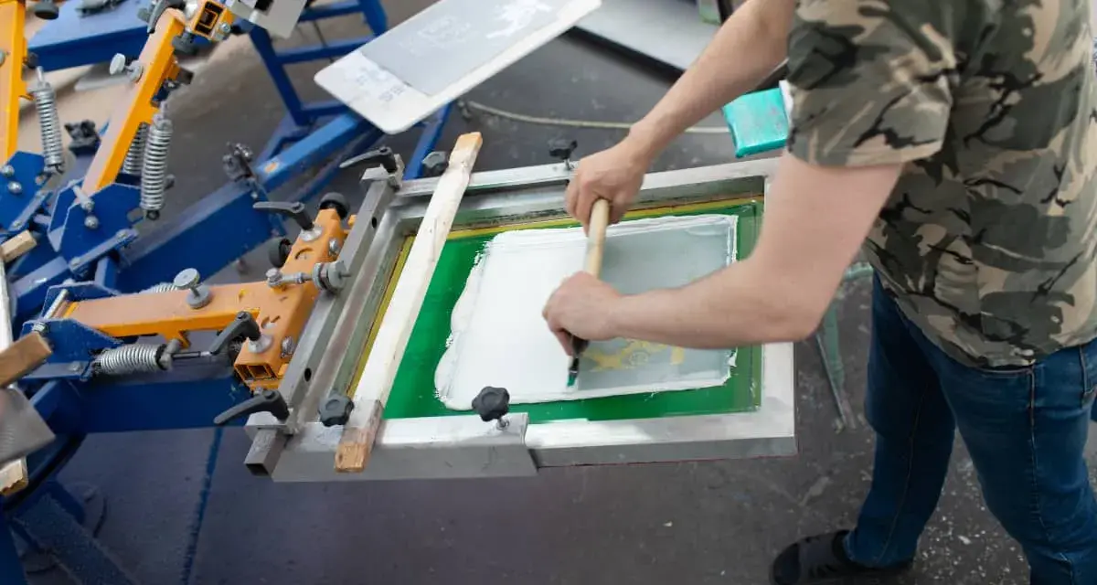 Proceso de serigrafía que es: un operario aplica tinta blanca sobre una pantalla para estampar.