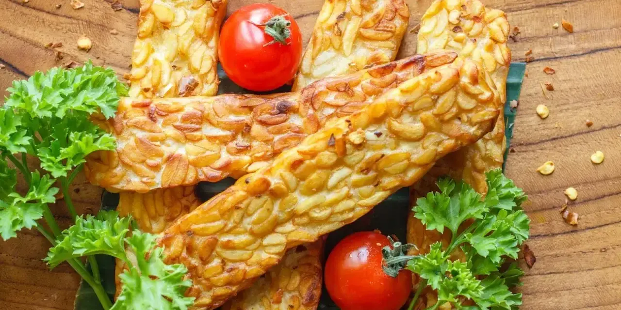 Złocisty, smażony tempeh, czyli co to jest? Podany z pomidorkami i natką pietruszki na drewnianej desce.