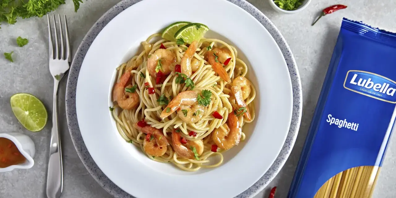 Pyszny makaron z krewetkami na winie, udekorowany limonką i natką pietruszki. Obok opakowanie makaronu spaghetti Lubella.