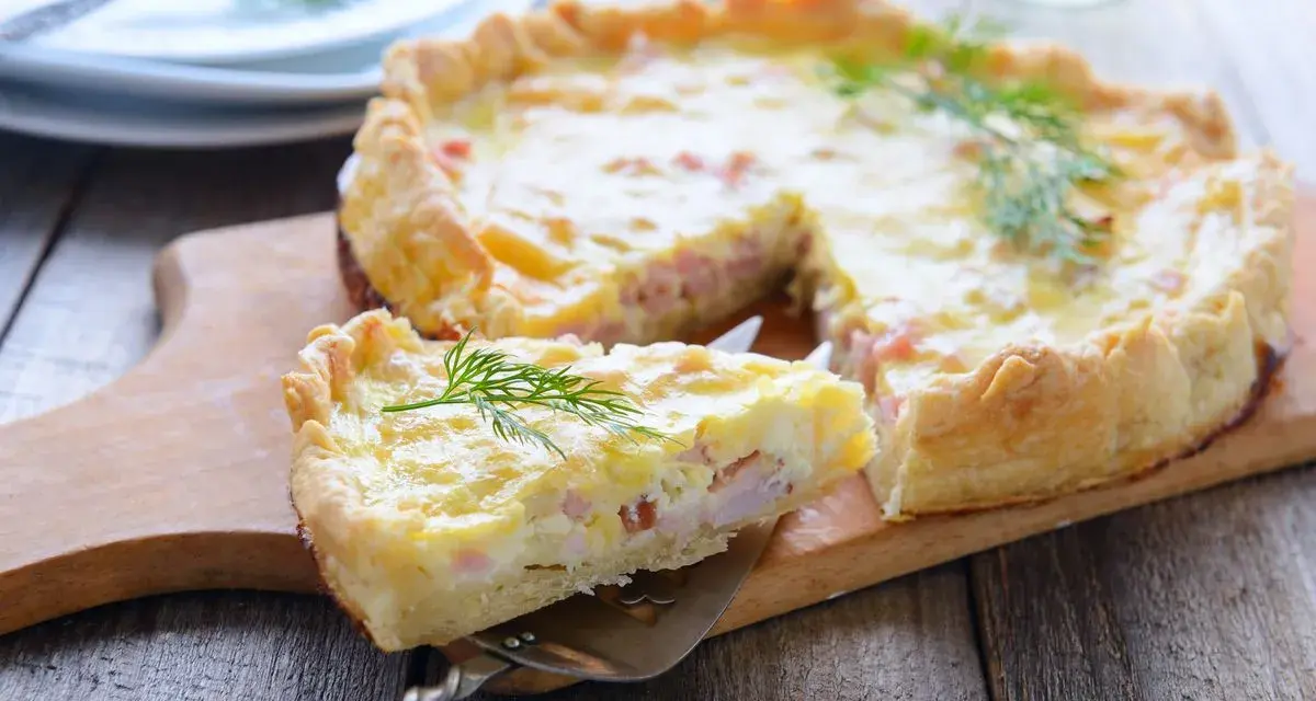 Una fetta di torta salata prosciutto e formaggio, guarnita con aneto, su un tagliere di legno.