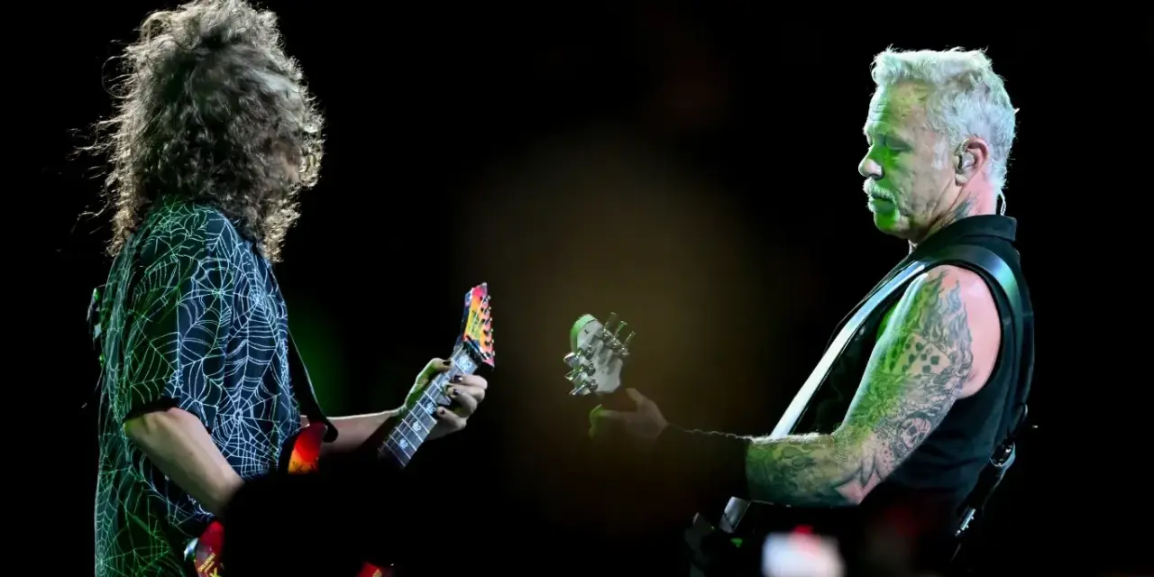 James Hetfield, z charakterystycznym wąsem i wytatuowanymi ramionami, gra na gitarze. Obok niego drugi muzyk z burzą włosów.