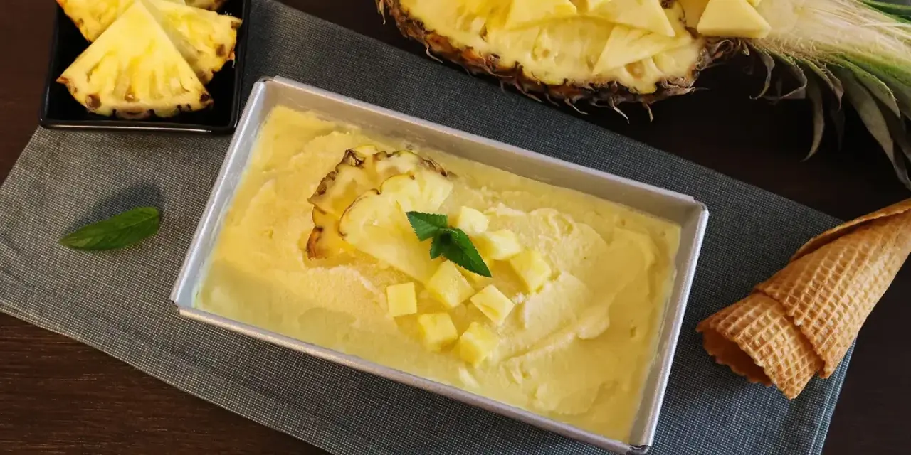 Erfrischendes Ananas Eis in einer Metallform, garniert mit Ananasstücken und Minze. Daneben liegen Ananasspalten und eine Waffel.